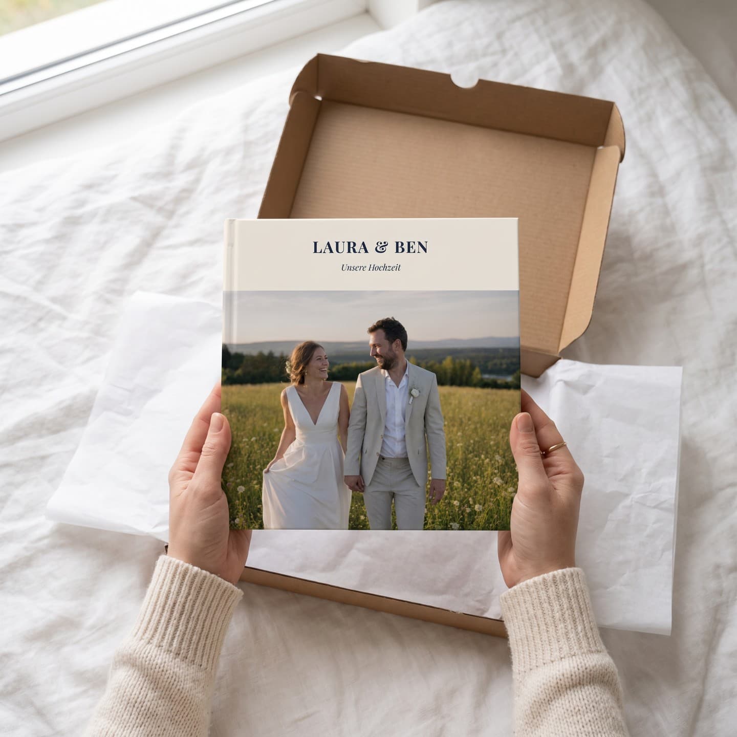 Zwei Hände heben das Hochzeitsalbum „Laura & Ben" aus einem Versandkarton auf dem Bett