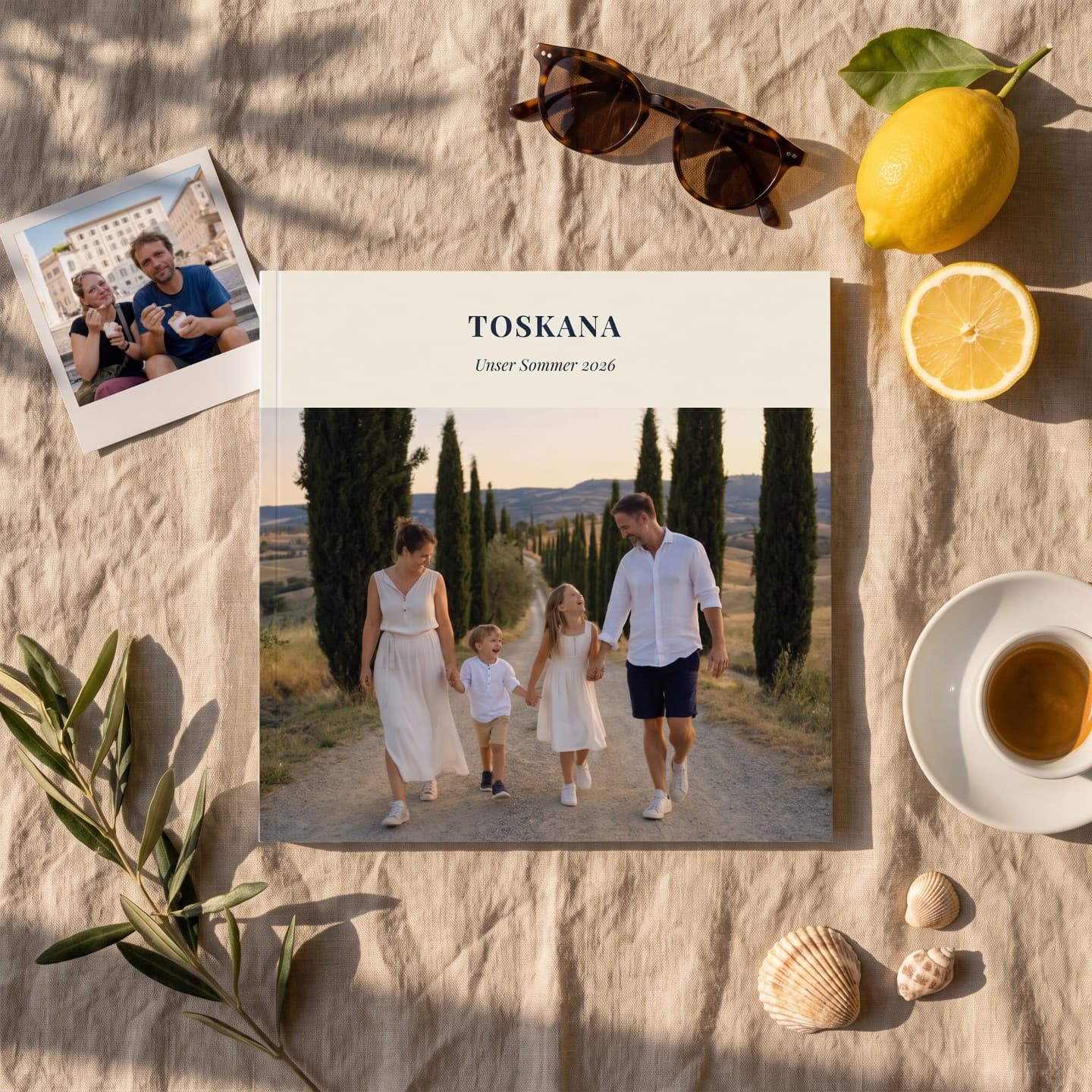 Reisefotobuch „Toskana" auf einer Leinendecke, daneben ein Sofortbild, Sonnenbrille, Zitronen und Espresso