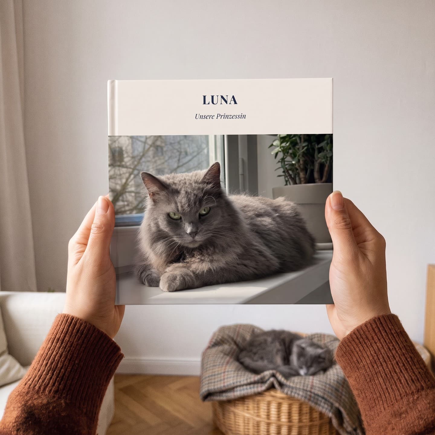 Fotobuch „Luna" in zwei Händen, dahinter schläft eine Katze im Korb